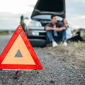 Broken car concept, man sitting on tire against breakdown triangle on asphalt road. Problem with vehicle, warning sign