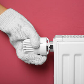Female hand in warm mitten on temperature regulator of heating battery on pink background, closeup