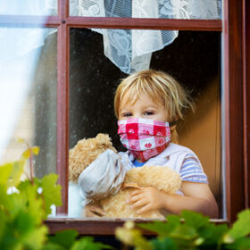 Sweet toddler boy, wearing medical mask, hugging teddy bear, also with mask, looking sadly out of the window, during coronavirus pandemic isolation