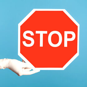 Profile side view closeup of human hand in white surgical gloves holding and showing Stop sign. indoor, studio shot, isolated on blue background.