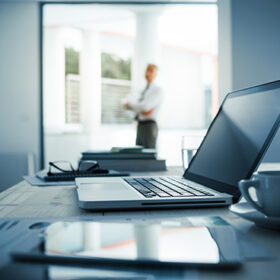 Businessman standing with arms crossed in his office, desktop with laptop on foreground, selective focus