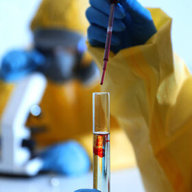 Scientist in chemical protective suit dripping blood into test tube at laboratory, closeup. Virus research